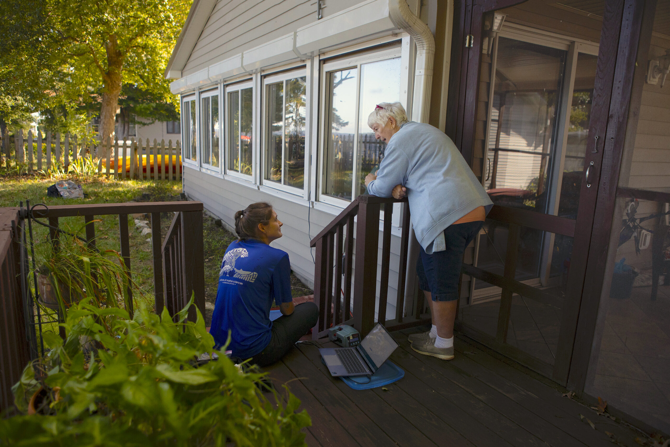 A woman leans against a porch railing and talks with a researcher, who is seated on the porch step.