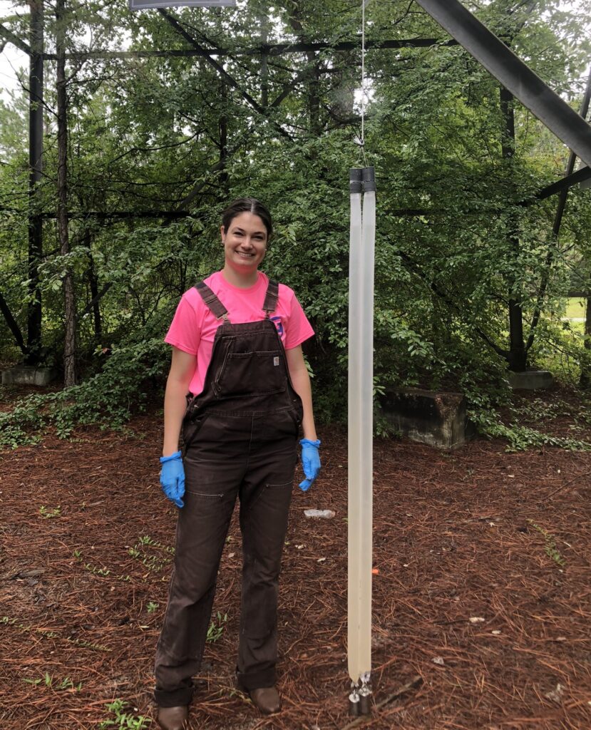 Tiffany VanDerwerker stands smiling alongside two Hydrasleeves, a long thin tube used for groundwater sampling, which contain water samples from the NCDEQ monitoring wells. 