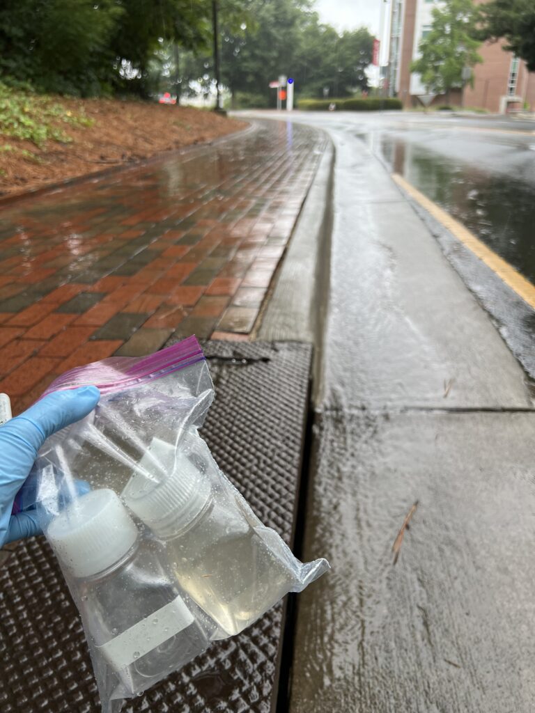View of a street in the background. Gloved hand in the foreground holding a plastic bag with water samples.