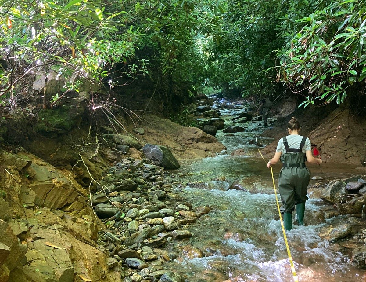 A person in waders walks through a rocky creek holding onto a rope